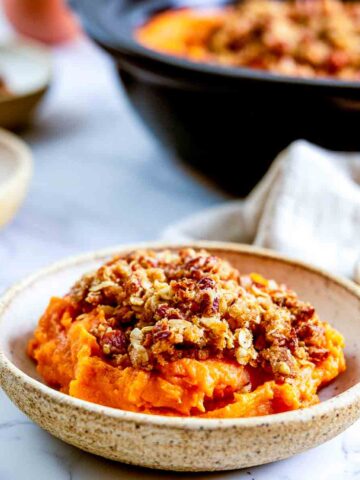 Close-up side view of a bowl of crockpot sweet potato casserole with golden pecan streusel on a white marble counter.