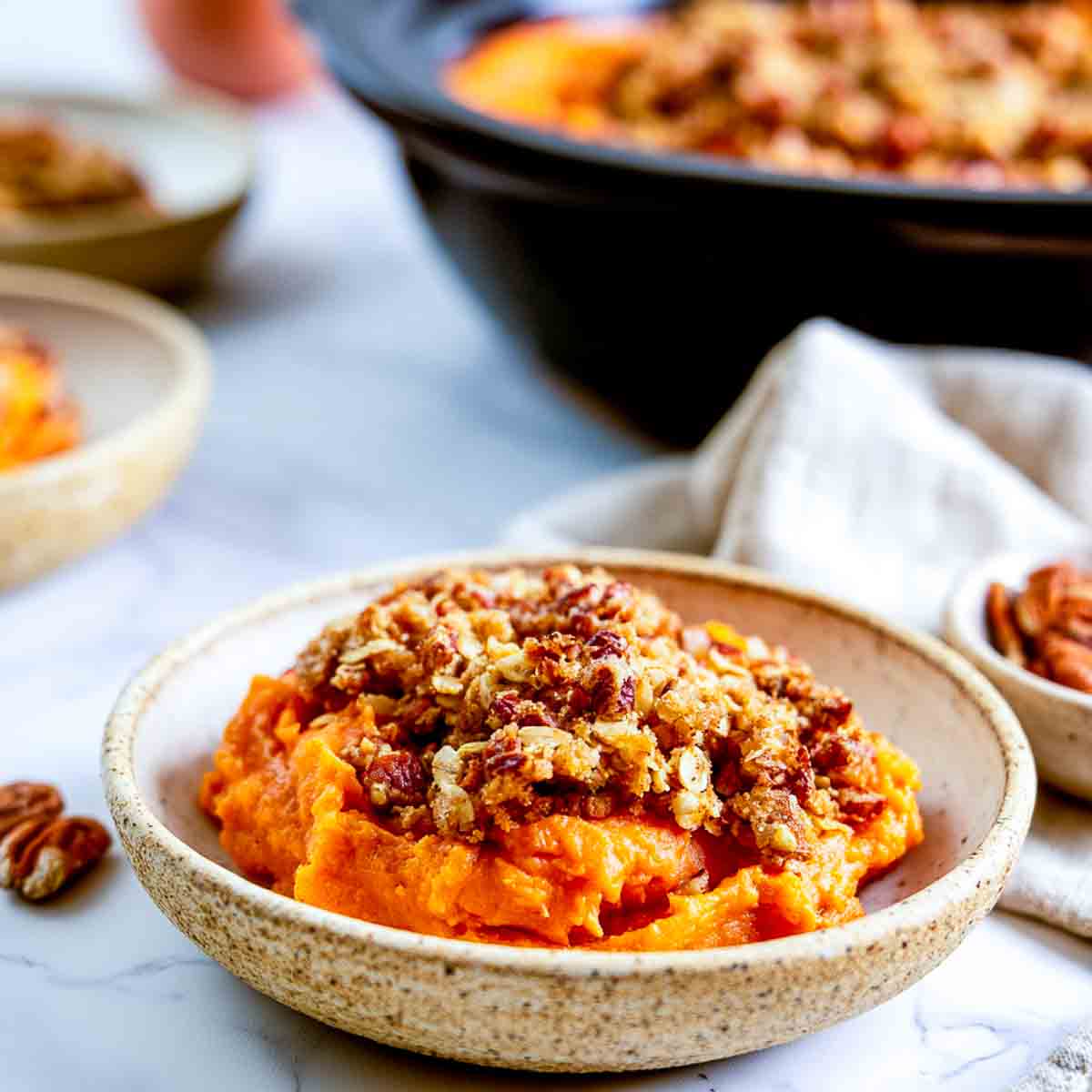 Close-up side view of a bowl of crockpot sweet potato casserole with golden pecan streusel on a white marble counter.