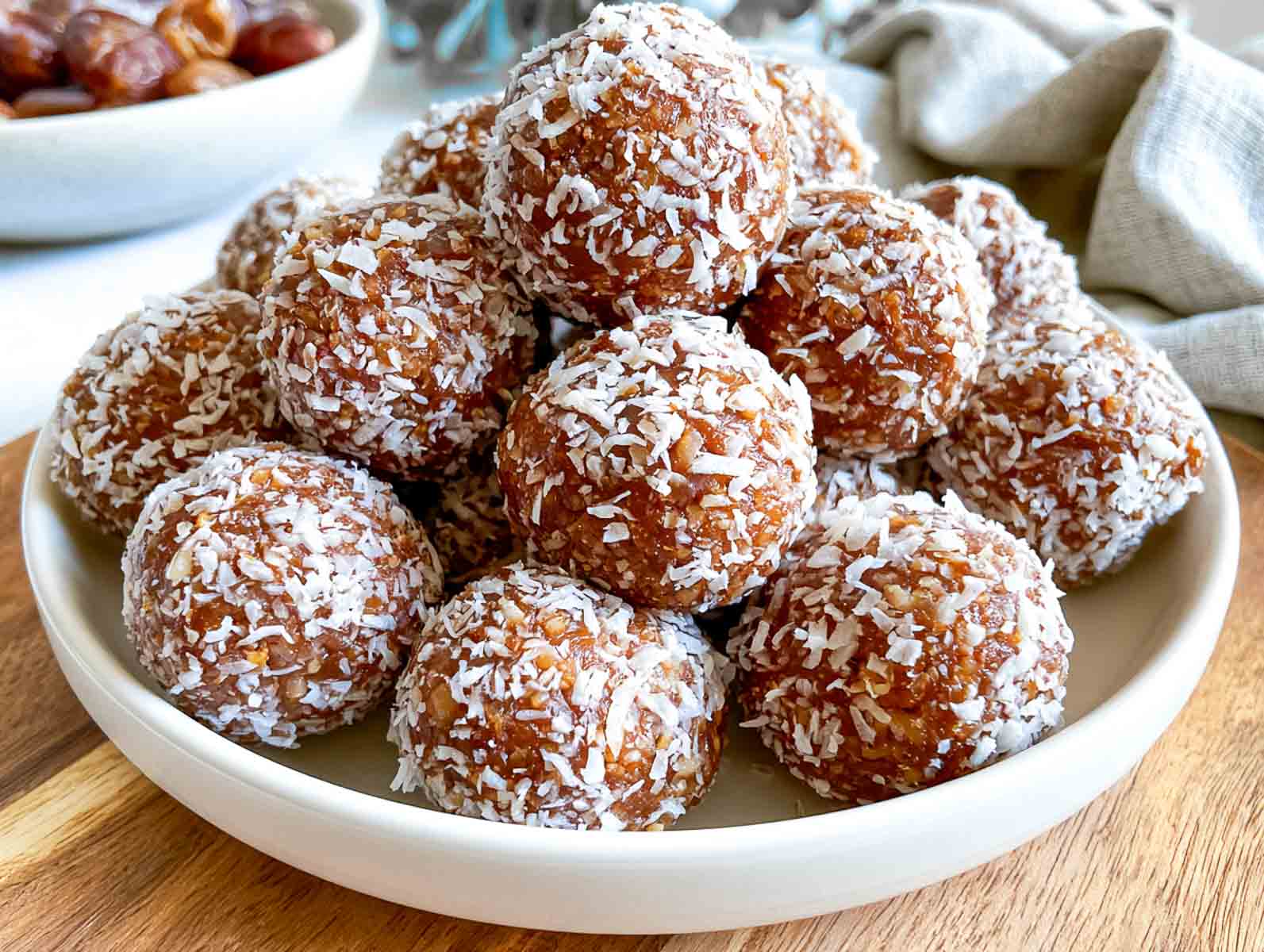 Vegan date-sweetened coconut high-protein energy balls rolled in shredded coconut on a plate, with a bowl of Medjool dates behind.