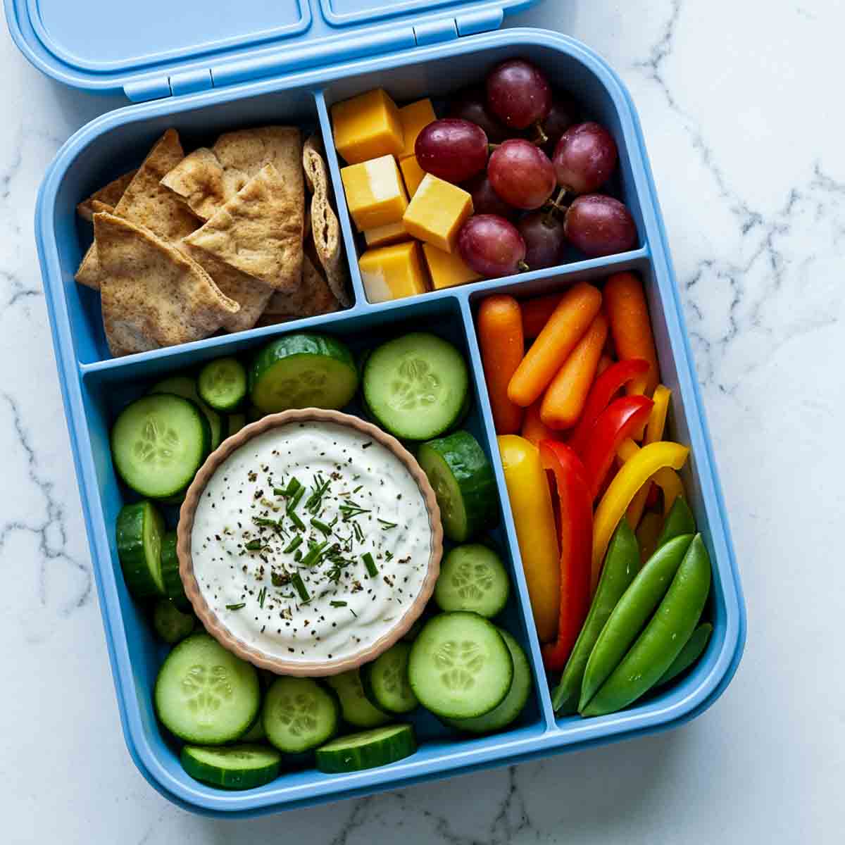 Blue lunchbox filled with cucumber slices and Greek yogurt ranch dip, pita chips, cheese cubes, red grapes, carrot sticks, bell pepper strips, and snap peas.