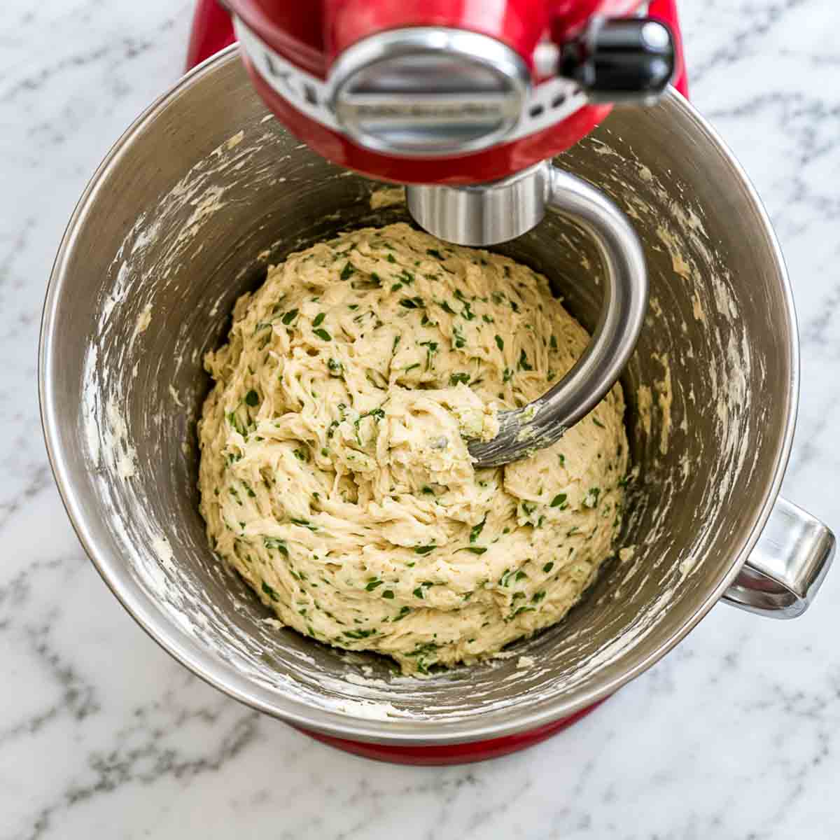 Shaggy kefir bread dough with chopped herbs coming together in a stand mixer bowl.