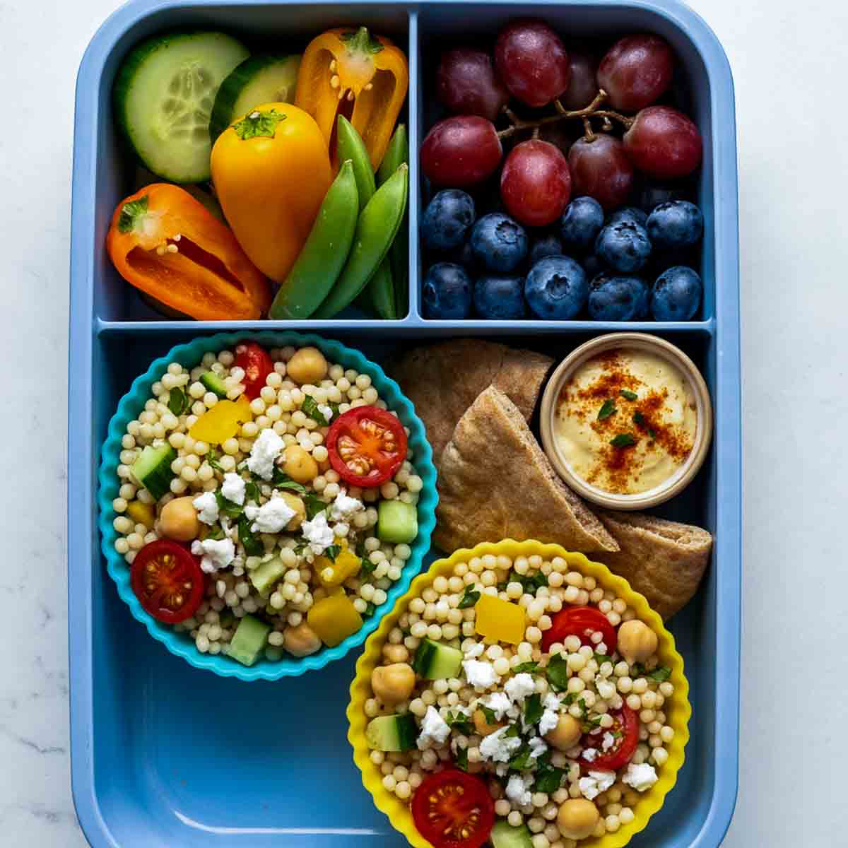 Blue lunchbox with Mediterranean couscous salad cups containing chickpeas, cucumber, tomatoes, peppers, and feta, served with pita chips and hummus.