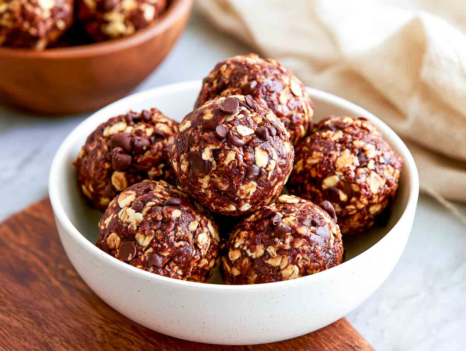 Nut-free chocolate oat high-protein energy balls topped with mini chocolate chips in a white bowl on a wooden board.