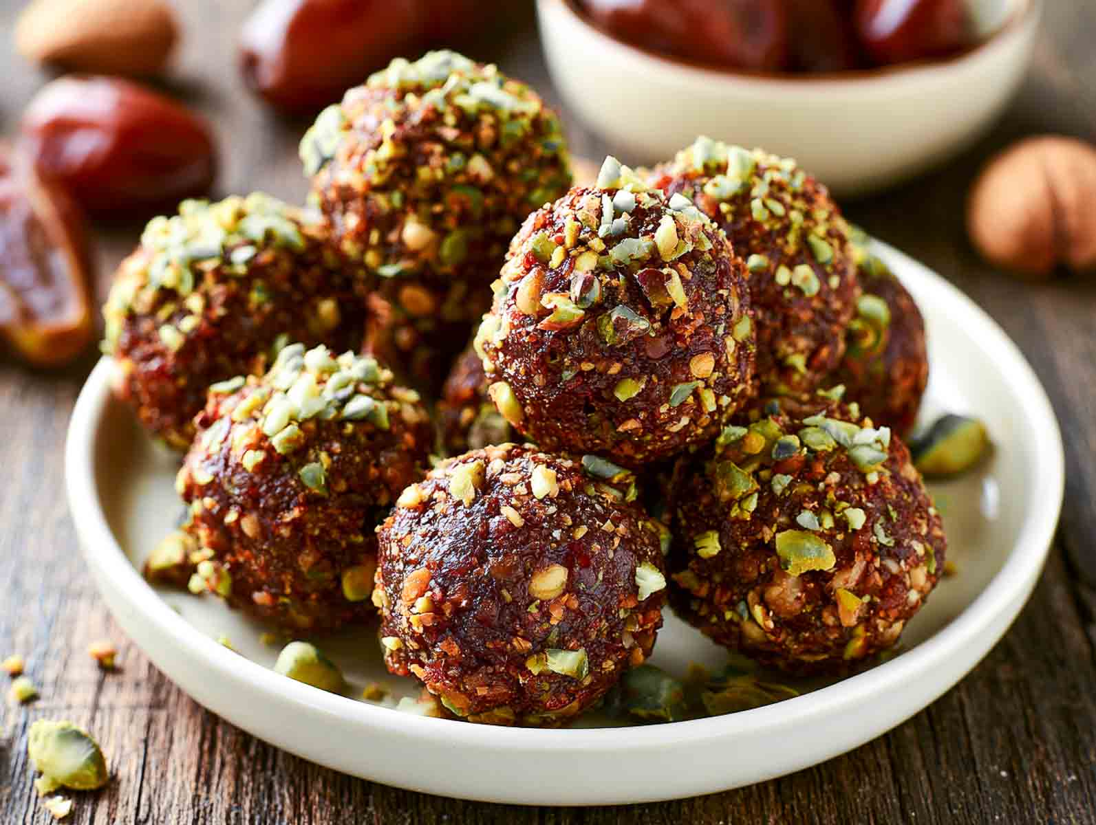 Pistachio date high-protein energy balls topped with chopped pistachios on a white plate, with Medjool dates and walnuts nearby.