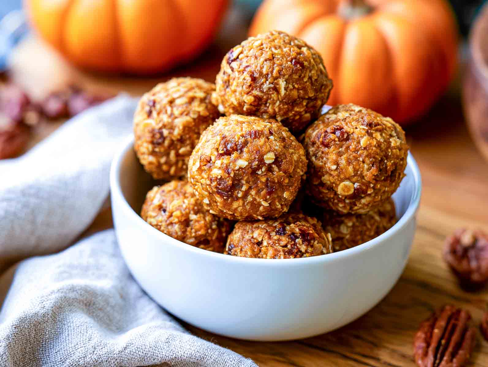 Pumpkin spice high-protein energy balls with oats, pecans, and dried cranberries in a white bowl, with small pumpkins and pecans nearby.