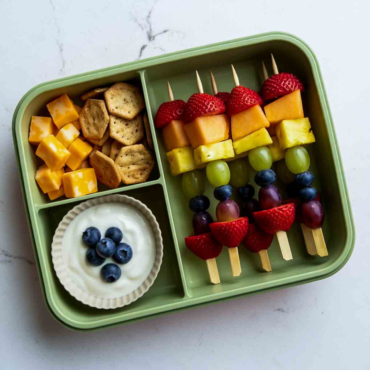 Green lunchbox with rainbow fruit kabobs made with strawberries, cantaloupe, pineapple, grapes, and blueberries, served with yogurt dip, cheese cubes, and crackers.