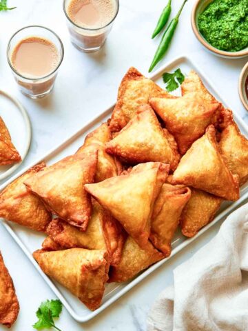 A rectangular white platter is piled with golden fried samosas, with two chutneys, two glasses of chai, green chilies, cilantro, and a beige napkin arranged around it.