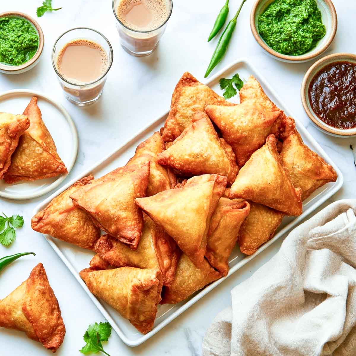 A rectangular white platter is piled with golden fried samosas, with two chutneys, two glasses of chai, green chilies, cilantro, and a beige napkin arranged around it.