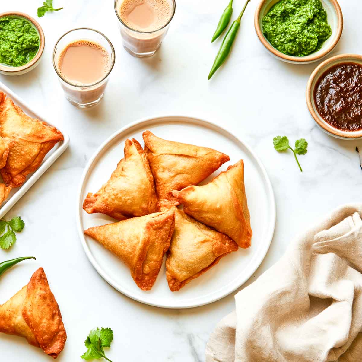 Five golden samosas are arranged on a round white plate, with two glasses of chai, bowls of chutney, green chilies, cilantro, and a folded beige napkin around the plate.