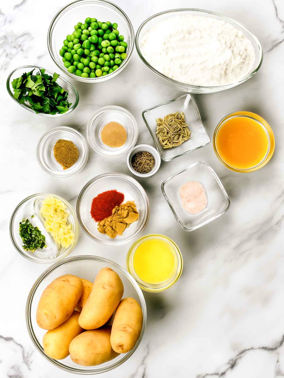 Ingredients for samosa in glass bowls on white marble, including potatoes, peas, flour, ghee, oil, cilantro, ginger, green chili, whole seeds, and ground spices.