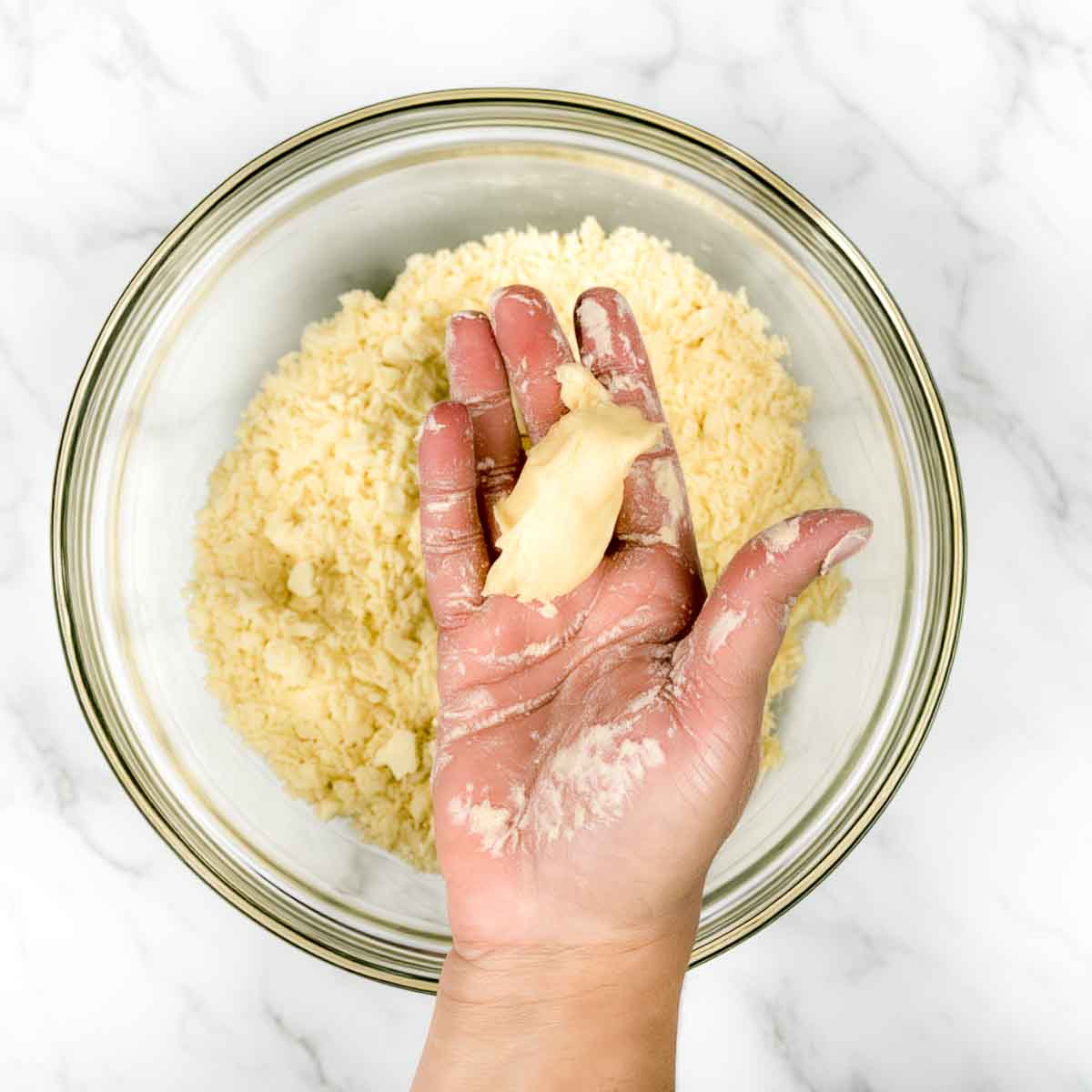 A hand holding a pressed clump of flour and ghee over a bowl, showing the samosa dough holding its shape.