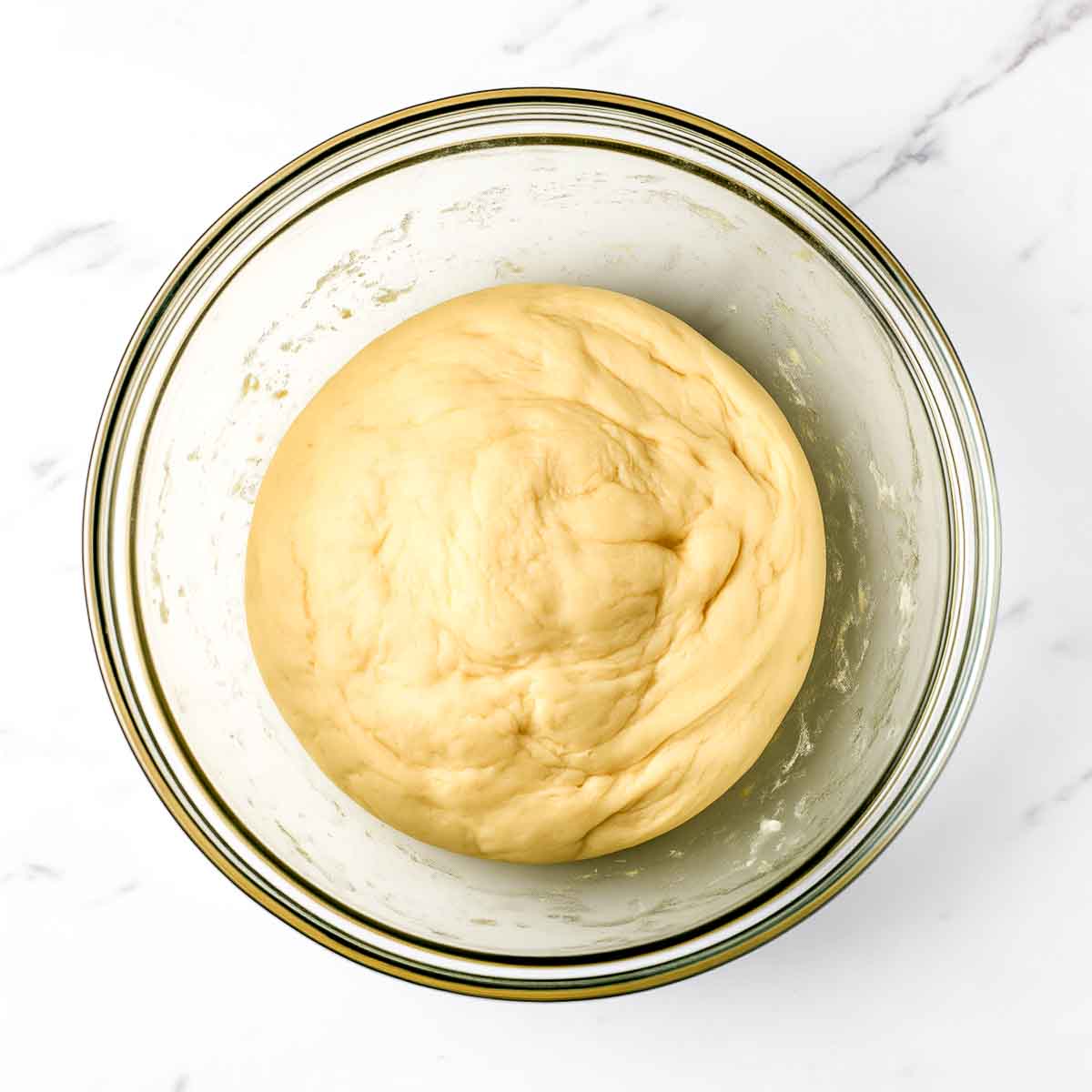 A smooth, firm ball of samosa dough resting in a glass bowl on white marble.