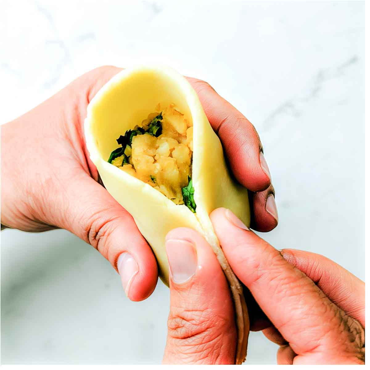Hands hold an open samosa cone filled with the seasoned potato mixture while the edges are being brought together for sealing.