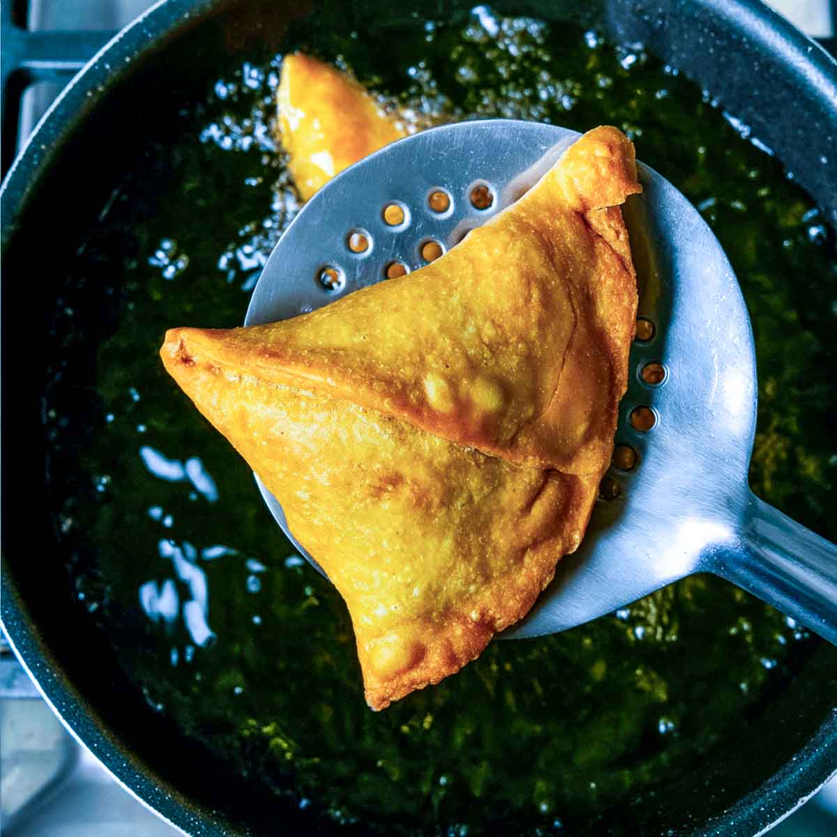 A golden samosa is lifted from hot oil with a slotted spoon as it finishes frying in a pan.