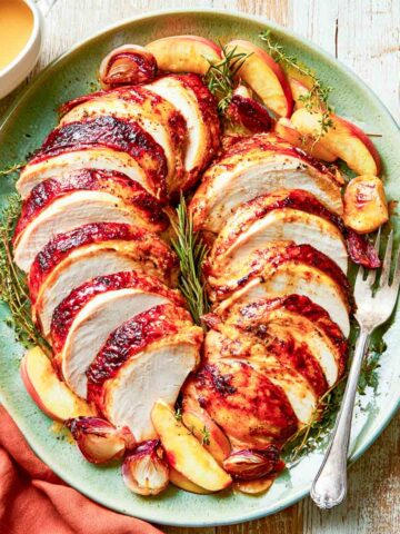 Sliced slow cooker turkey breast with maple Dijon glazed skin on a sage green platter with roasted apples, shallots, thyme, gravy, and cranberry sauce.