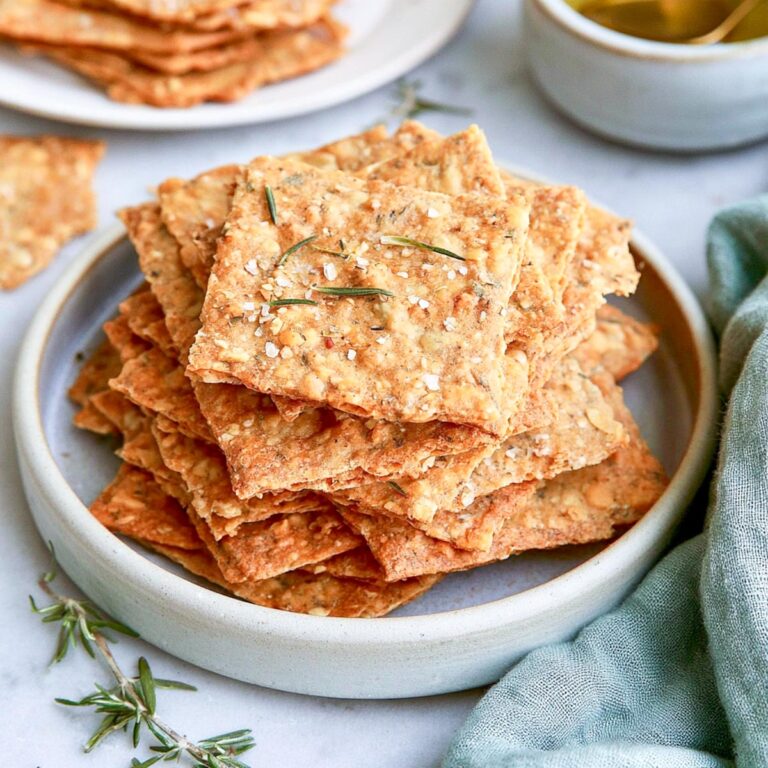 Close-up of crisp, golden sourdough crackers stacked on a ceramic plate with rosemary and a green linen napkin.