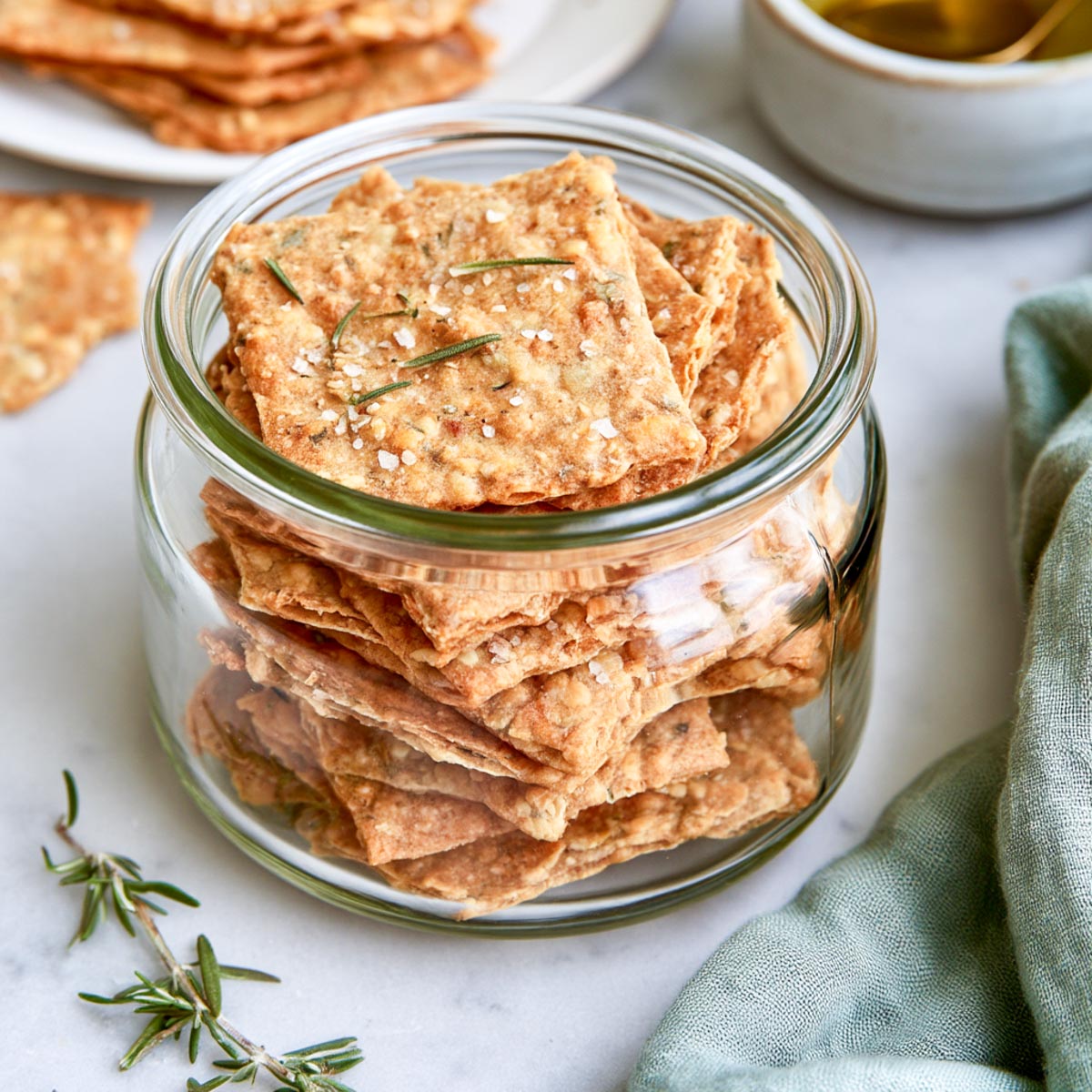 Sourdough crackers stacked in a clear glass storage jar with rosemary and a green linen napkin on white marble.