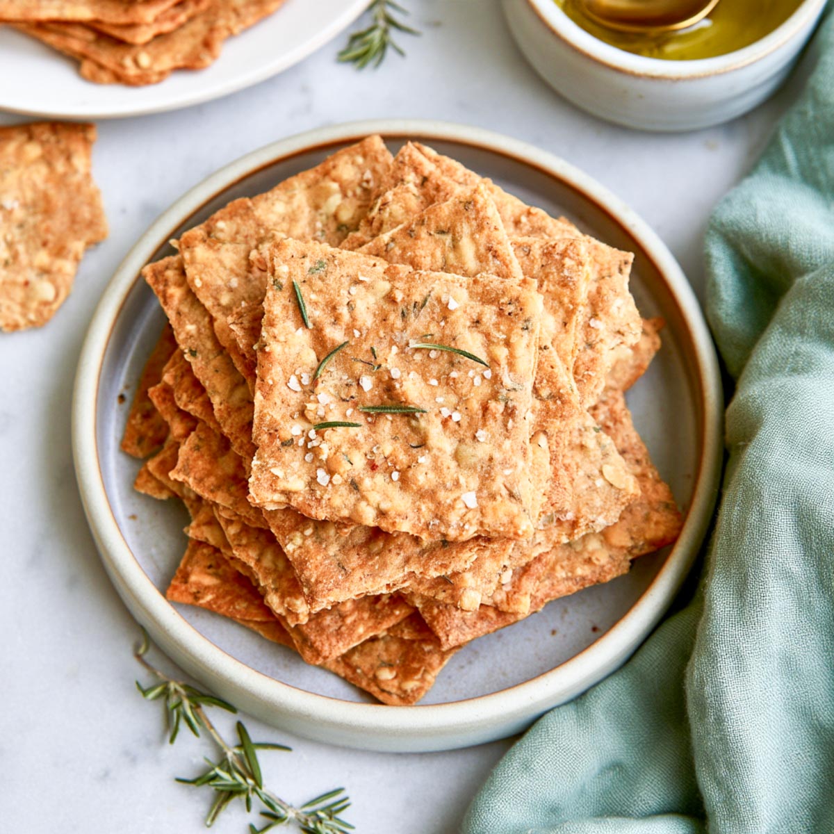 Top-down view of golden sourdough crackers stacked on a ceramic plate with rosemary and a green linen napkin.