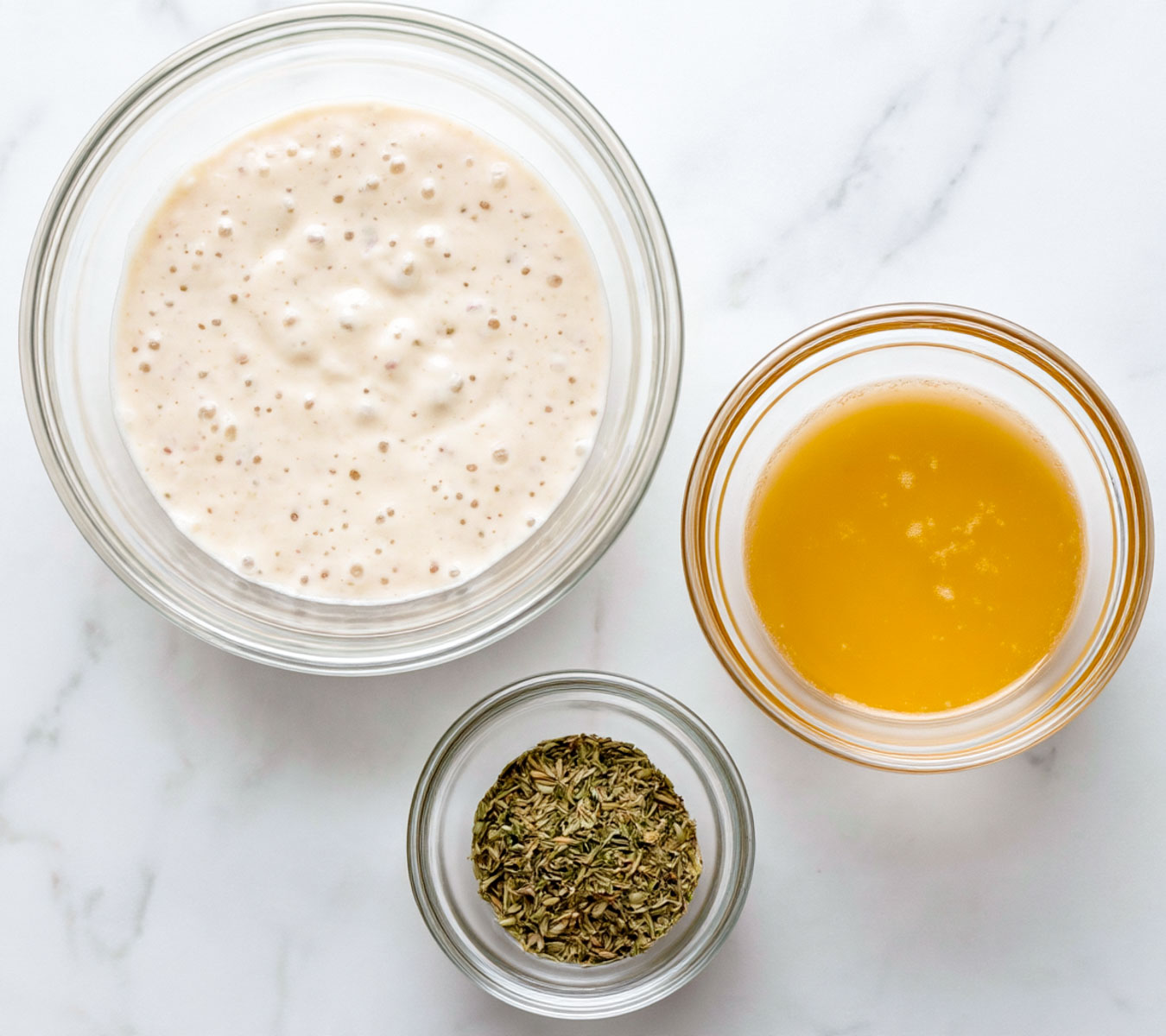 Sourdough cracker ingredients arranged on white marble, including sourdough discard, melted butter, and dried herbs in glass bowls.
