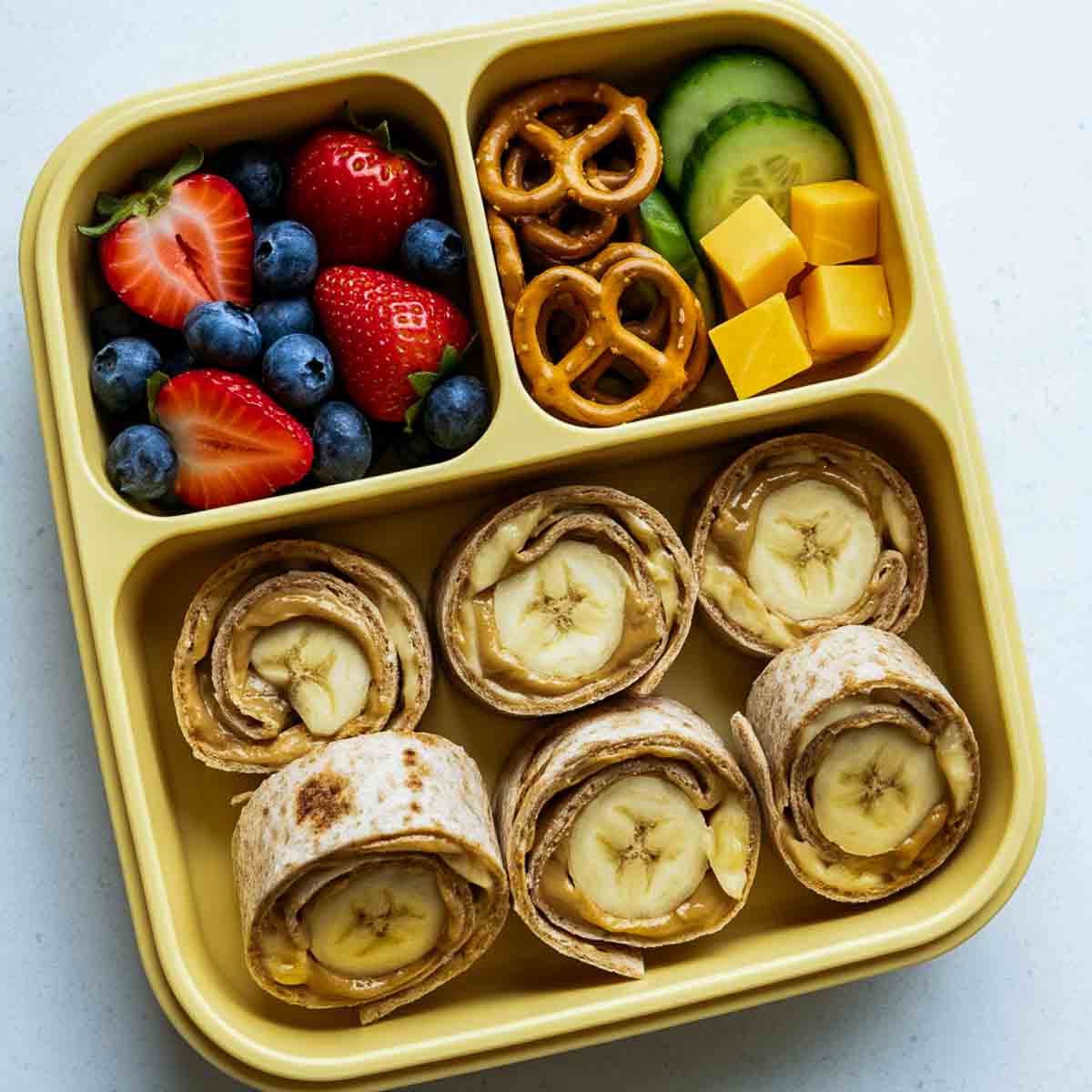 Yellow lunchbox filled with sunbutter banana roll-ups, strawberries and blueberries, pretzels, cucumber slices, and cheddar cheese cubes.