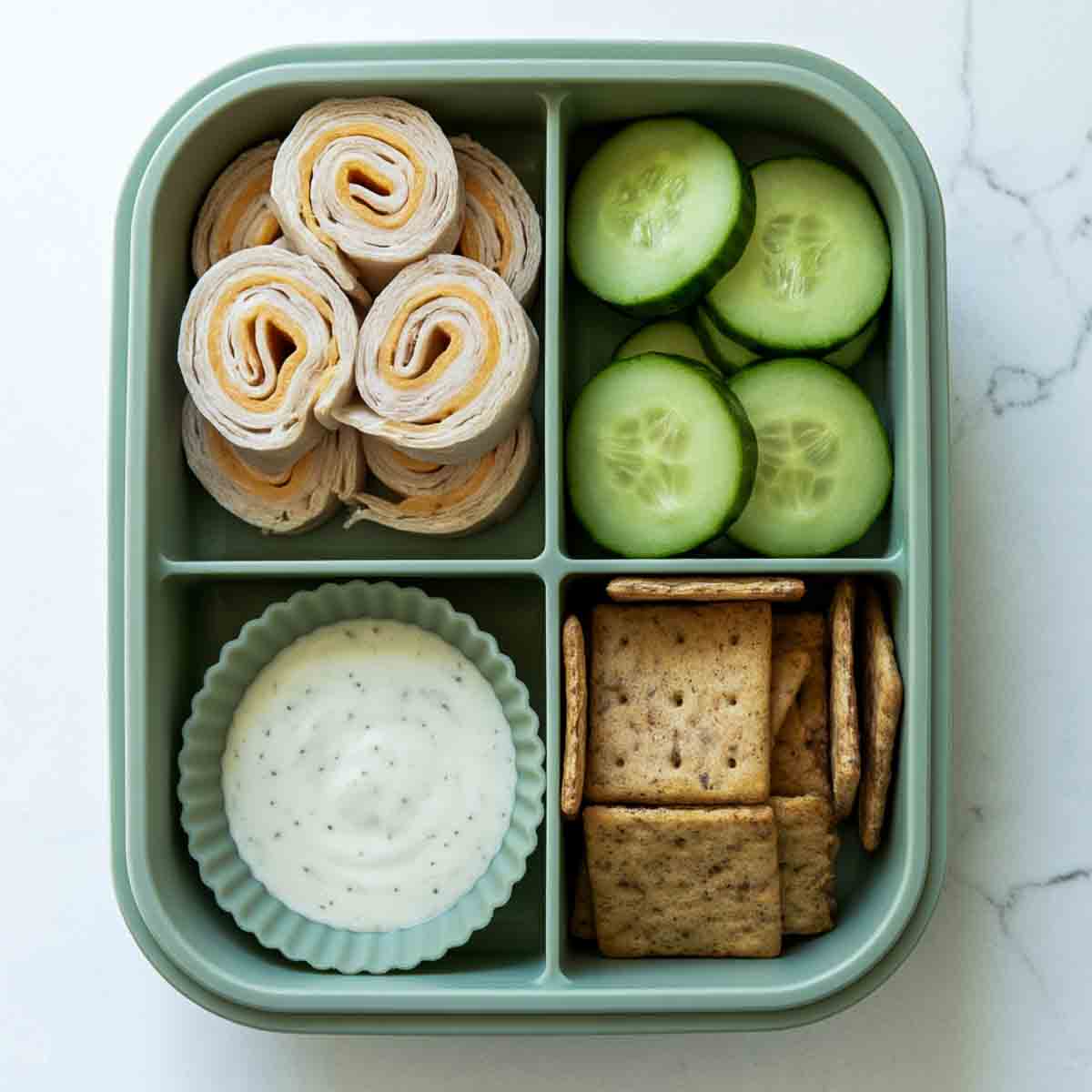 Green lunchbox with turkey and cheese roll-ups, cucumber slices, ranch dip, and whole-grain crackers.