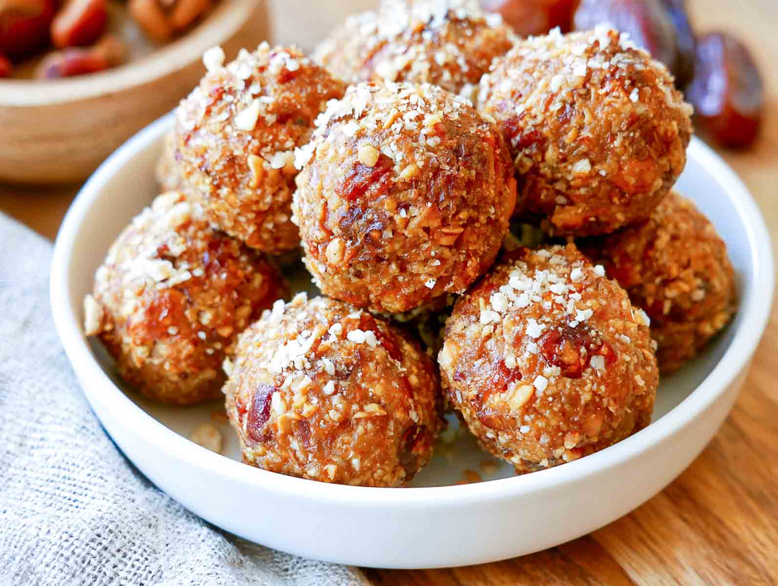 Vegan peanut butter date high-protein energy balls rolled in crushed peanuts and sprinkled with coconut in a white bowl, with Medjool dates behind.