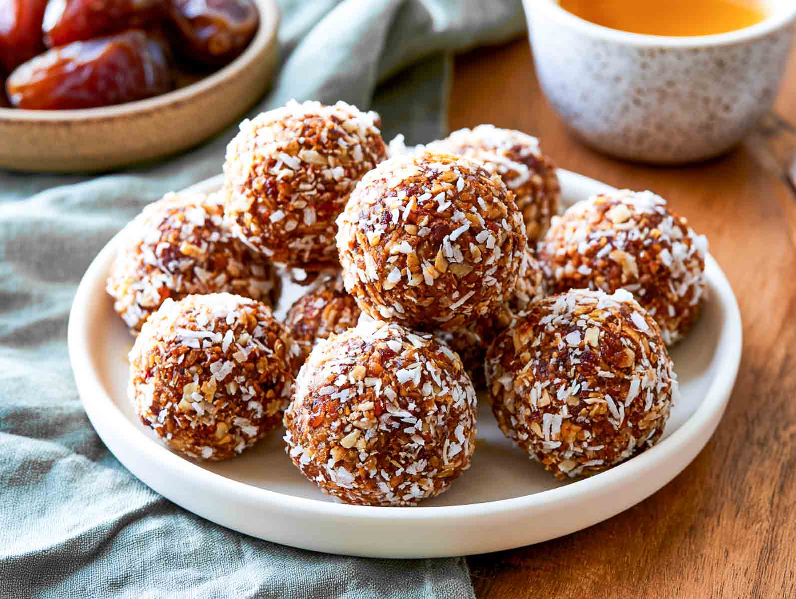 Whole30 date coconut high-protein energy balls rolled in shredded coconut on a white plate, with Medjool dates and a cup of tea behind.